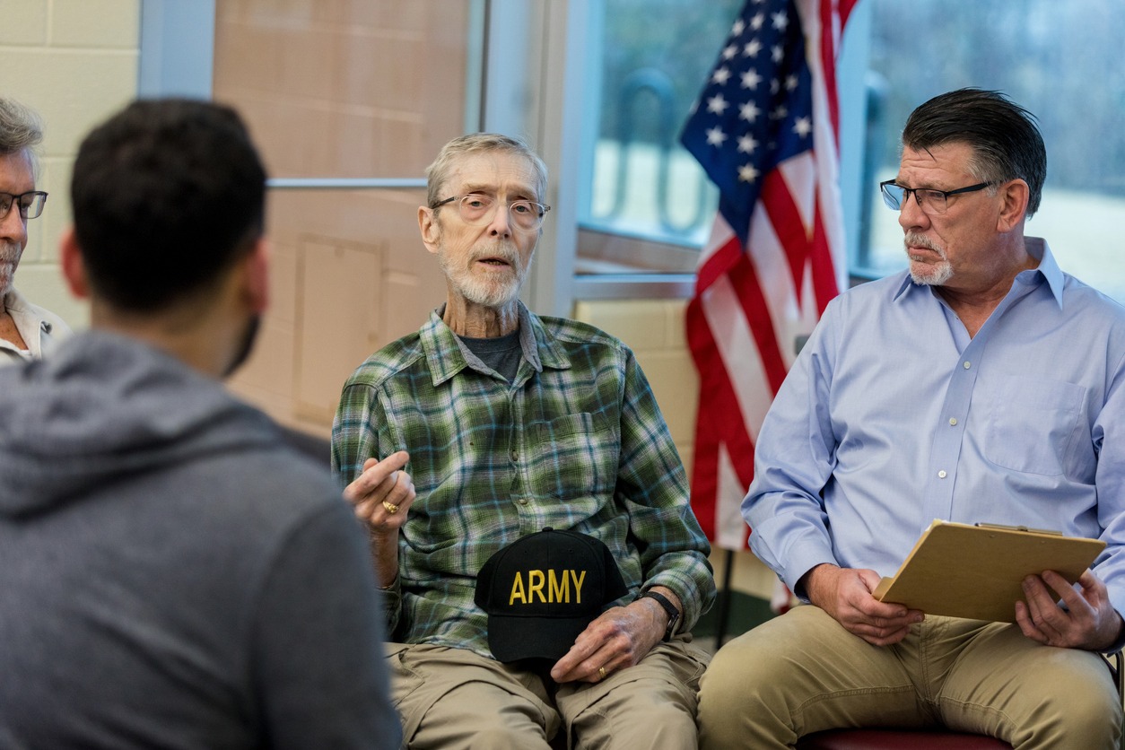 Older man in a plaid shirt speaks during a veterans' discussion, holding an Army cap; an American flag is in the background.