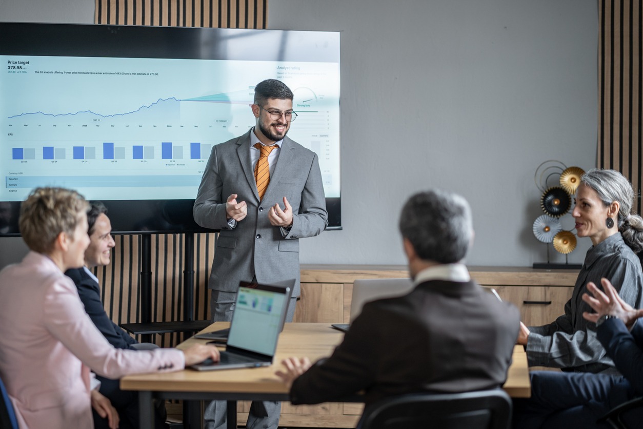 Man in a gray suit and orange tie presenting financial charts to colleagues in a meeting room.