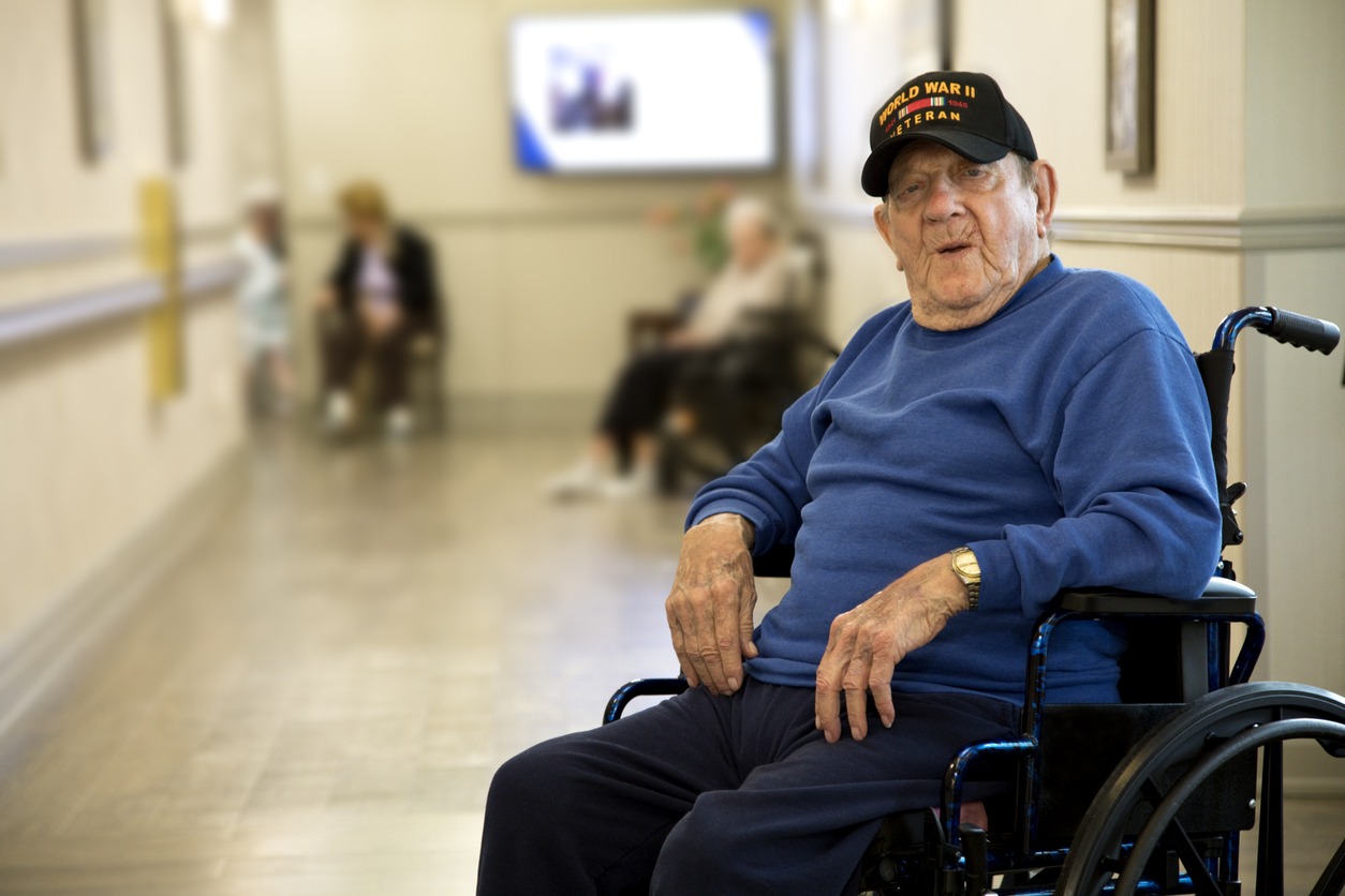 Older man in a wheelchair in a hospital corridor, wearing a blue sweater and a World War II veteran cap.